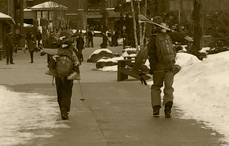 An image of Ty and Dylan walking through the Spruce Peak Village with their skis and gear at Stowe Mountain Resort in Vermont