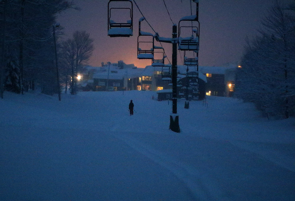 An image of Ty finishing a ski tour at dusk with the glow of the Bolton Valley Village behind him at Bolton Valley Resort in Vermont