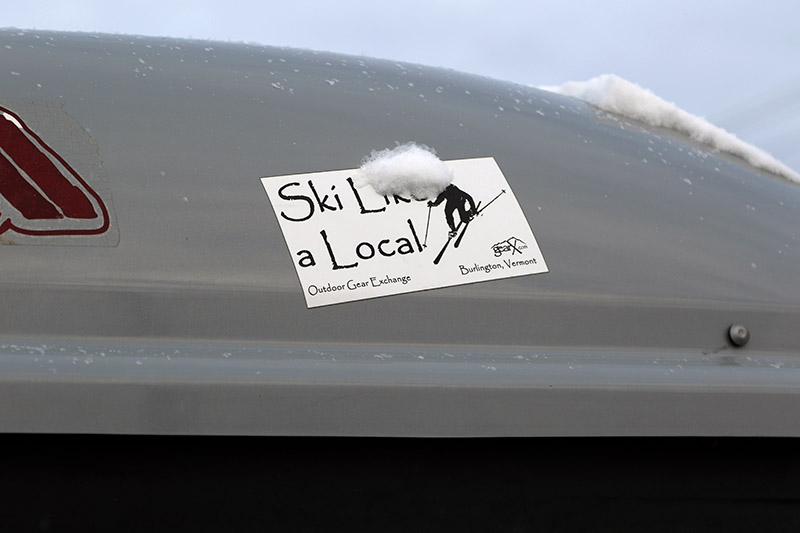 An image showing the logo for the Outdoor Gear Exchange store on a roof-top ski box of a nearby car while skiing at Bolton Valley Resort in Vermont