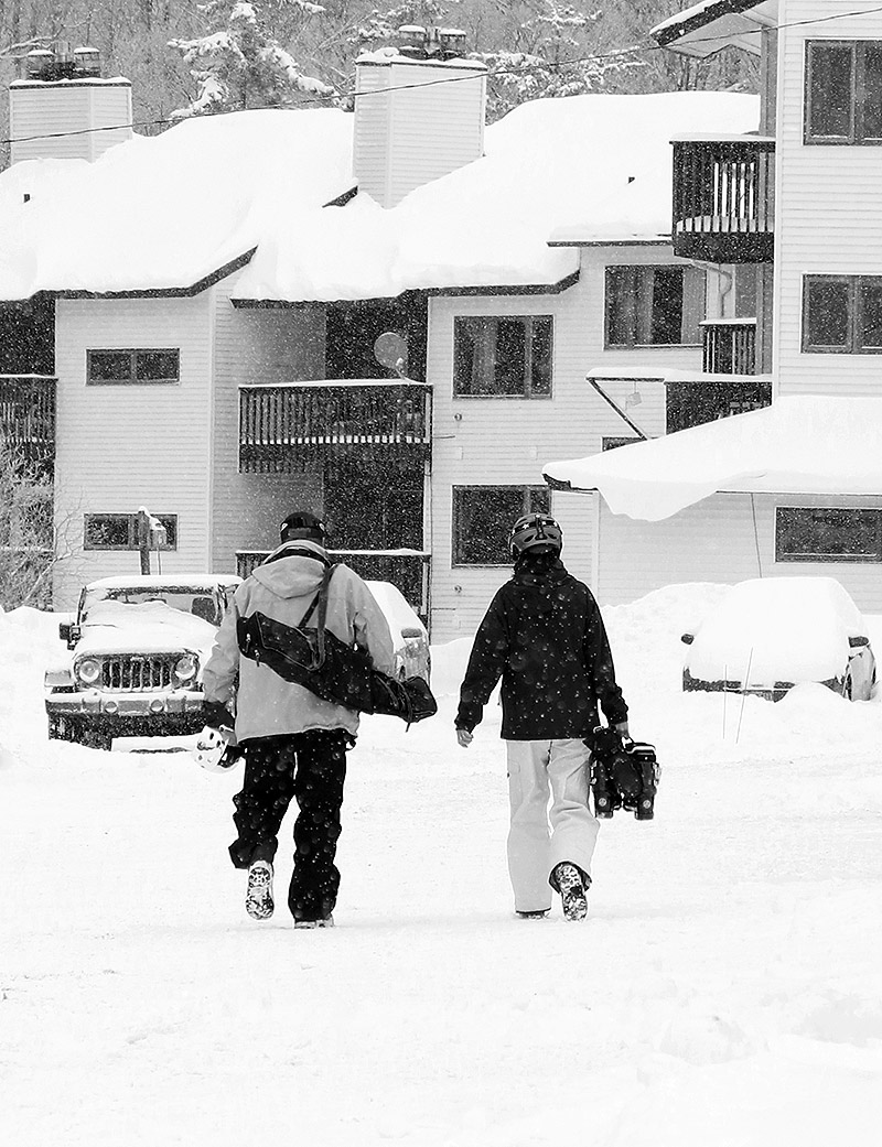 An image of two skiers walking through snowfall in the Village at Bolton Valley Ski Resort in Vermont