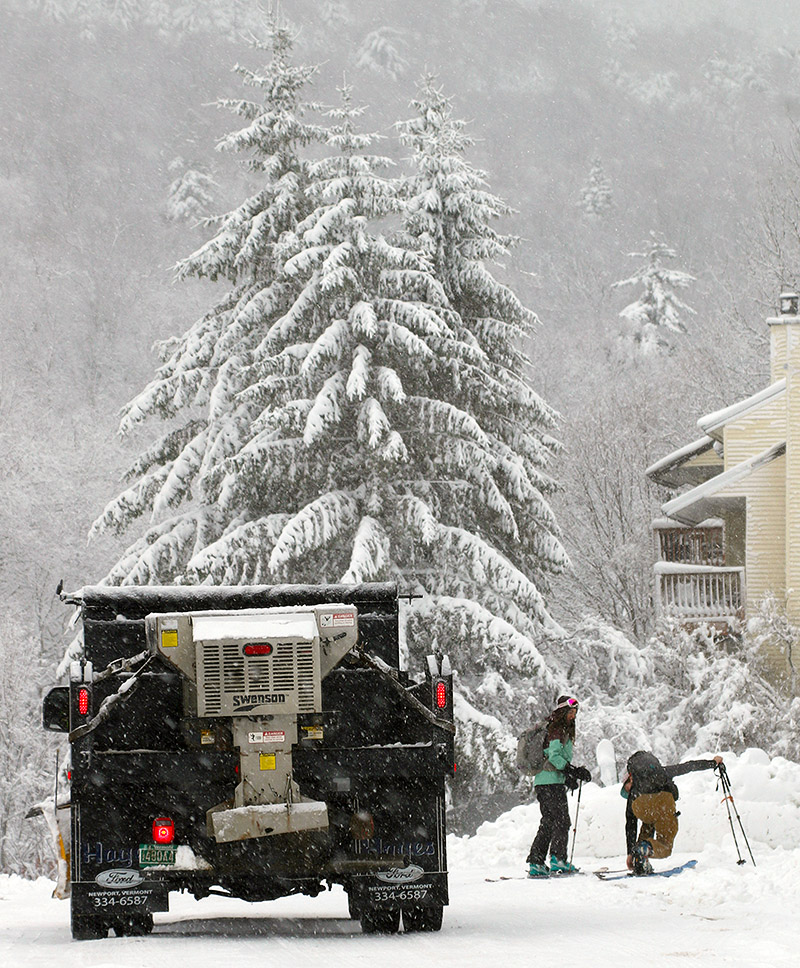 An image of skiers getting ready for a ski tour and a snow plow working to maintain roads during an early December snowstorm in the Village area of Bolton Valley Ski Resort in Vermont