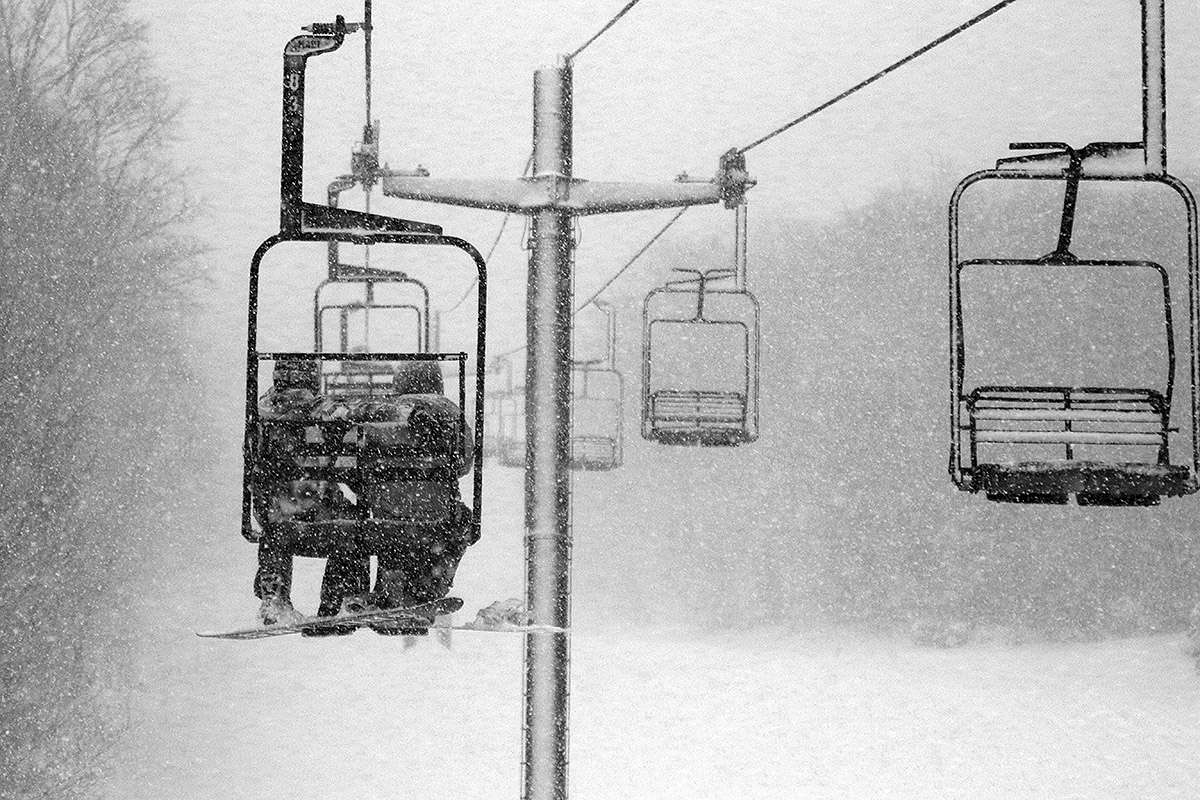 An image of two snowboarders riding the Wilderness Double Chairlift as heavy snowfall in the 1 to 2-inch per hour range falls at Bolton Valley Resort in Vermont during Winter Storm Gerri
