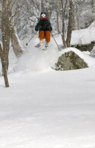 An image of Ty on Telemark skis jumping off a rock into deep powder in the Moose Glen area of the Nordic and Backcountry Network during a February ski tour at Bolton Valley Ski Resort in Vermont