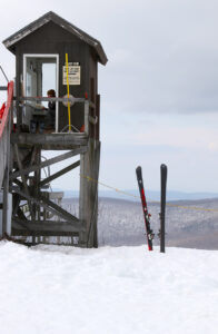 An image of the Mid Station area of the Timberline Quad Chairlift with a lift attendant and a pair of skis stuck in the snow on a warm February day at Bolton Valley Ski Resort in Vermont