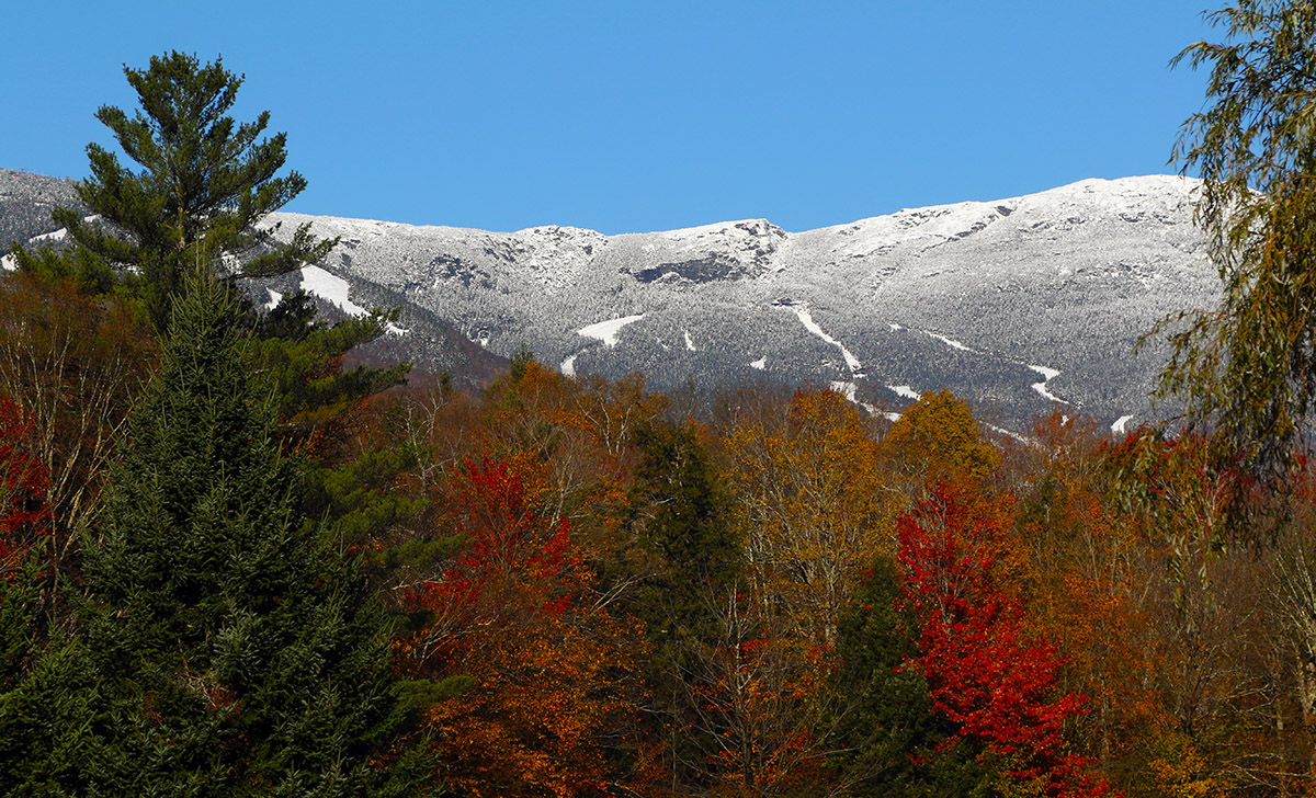 A view of some of the snowy trails of Stowe Mountain Resort in Vermont from the Mountain Road under brilliant sunshine with foliage after a mid-October snowstorm