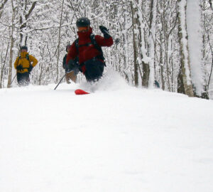 An image of Dylan Telemark skiing in the powder in the trees with friends looking on during a January ski tour in the Gilpin Mountain/Domey's Dome area of the Jay Peak backcountry in Vermont